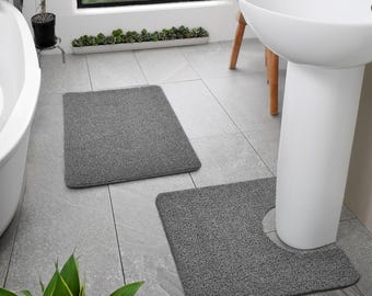 May include: Two gray bathroom mats. One is rectangular, and the other has a U-shaped cutout to fit around a pedestal sink. The mats are made of a textured material and are placed on a gray tiled floor.