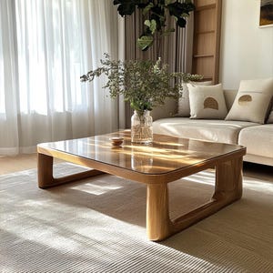 May include: A wooden coffee table with a glass top and rounded legs. The table is in a living room setting with a rug and a sofa in the background.