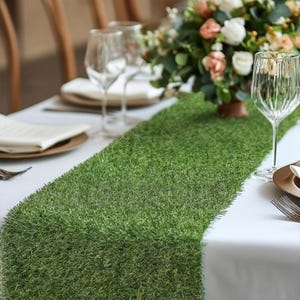 May include: A green artificial grass table runner on a white tablecloth with a vase of flowers and two wine glasses.