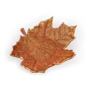 Puede incluir: Un plato decorativo en forma de hoja en tonos marrón y dorado. El plato tiene una superficie texturizada con vetas detalladas, que se asemejan a una hoja de arce. Los bordes están resaltados con dorado, añadiendo un toque de elegancia.