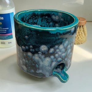 May include: A cylindrical ceramic vessel featuring a deep teal and dark blue glaze with a mottled, crystalline texture. The container has a small, protruding spout near the base, allowing for drainage, and a smooth, rounded rim at the top opening.