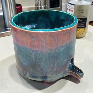 May include: A cylindrical ceramic mug featuring a reactive glaze with blended tones of teal, deep blue, and warm peach. The interior is a vibrant teal, and the exterior displays a smooth, glossy finish with a small, protruding spout at the base.
