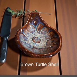 May include: A ceramic spoon rest with a pear-shaped design featuring a glossy brown and blue mottled glaze. The piece has three small holes at the top and is labeled Brown Turtle Shell. A black kitchen knife and a sprig of rosemary are nearby.