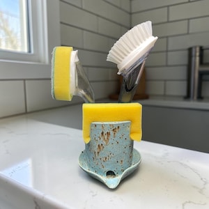 May include: A light blue ceramic sponge holder with brown speckles, featuring a wide rim and a small drainage spout at the base. A bright yellow rectangular sponge is draped over the top edge, while a dish brush with white bristles sits inside.