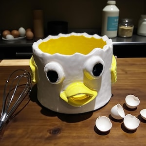 May include: A whimsical, white ceramic bowl with a yellow interior, resembling a cracked egg with a face. The bowl has large black eyes and a yellow beak. Eggshells are scattered around the bowl on a wooden surface.