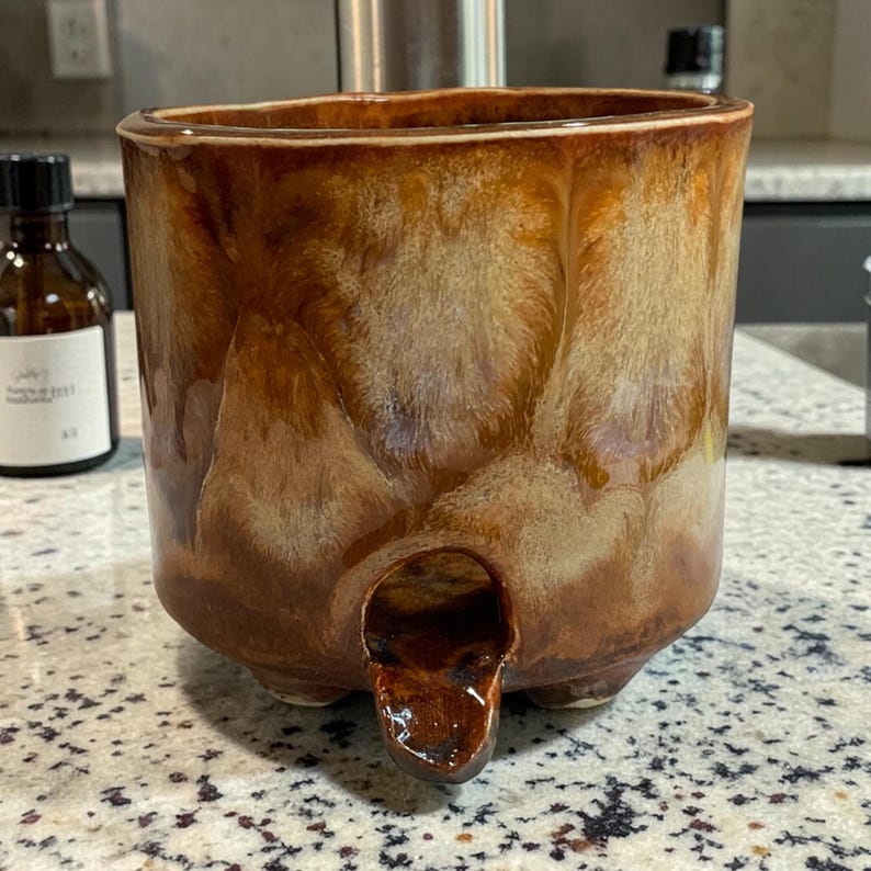 May include: A cylindrical ceramic planter with a glossy, variegated brown and cream drip glaze finish. The vessel features three small feet at the base and a prominent, arched drainage spout extending from the lower front section of the exterior wall.