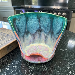 May include: A wide-mouthed ceramic bowl featuring a vibrant glaze that transitions from deep teal and blue at the top rim to a soft yellow and pink at the base. The interior surface displays a mottled, flowing texture with dark drips.