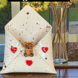 May include: Cream-colored ceramic envelope-shaped wall decor with a brown teddy bear and three red heart accents. The text "Bright Smiles!" is displayed on a black background above the envelope.