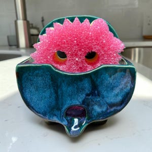 May include: A deep teal ceramic sponge holder with a glossy, variegated finish and a small drainage spout at the bottom. Inside the holder sits a bright pink, circular, textured scrubbing sponge with two distinct dark circular cutouts.