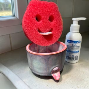 May include: A red, smiling sponge sits atop a gray and pink ceramic holder. A white bottle with a pump dispenser is to the right. The sponge has a face-like design, adding a playful touch to the kitchen accessory.