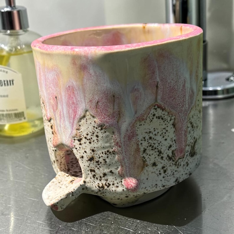 May include: A ceramic mug with a speckled cream base and a pink glaze dripping from the rim. The mug features a textured surface and a small, protruding spout-like element near the base, creating a distinct silhouette for this functional vessel.