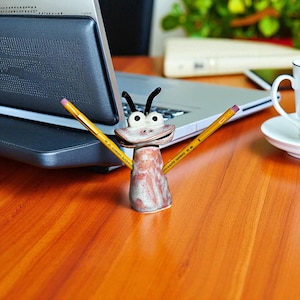 May include: A whimsical ceramic pencil holder shaped like a smiling bug with black antennae and white eyes. Two yellow pencils extend from the holder. The holder is on a wooden desk with a laptop and a coffee cup in the background.