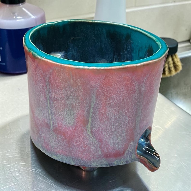 May include: A cylindrical ceramic mug featuring a vibrant teal interior glaze that drips over the rim. The exterior displays a soft, blended gradient of dusty pink and muted grey tones, with a small, dark teal spout protruding from the base.