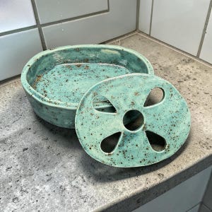 May include: An oval ceramic soap dish with a speckled light blue-green glaze. The dish consists of a deep base container and a removable lid featuring five circular and petal-shaped cutouts to allow for drainage and airflow for the soap.