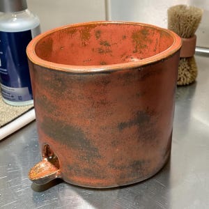 May include: A cylindrical ceramic vessel with a reddish-brown glaze featuring dark, mottled patches and a smooth, rounded rim. A small, protruding spout is attached to the lower side of the exterior wall, extending outward from the base.