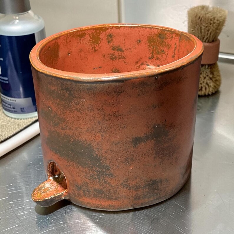 May include: A cylindrical ceramic vessel with a reddish-brown glaze featuring dark, mottled patches and a smooth, rounded rim. A small, protruding spout is attached to the lower side of the exterior wall, extending outward from the base.