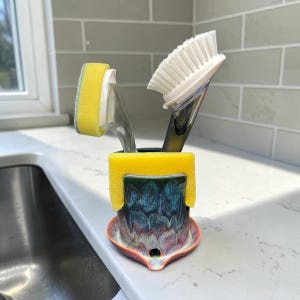 May include: A ceramic sponge holder with a multi-colored glaze in shades of blue, purple, and orange, featuring a small drainage hole at the base. A yellow rectangular sponge is draped over the rim, while a dish brush and a sponge wand sit inside.