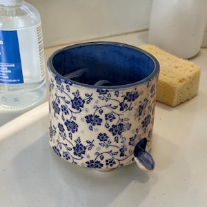 May include: A cylindrical ceramic shaving mug featuring a cream-colored base with a repeating dark blue floral pattern. The interior is glazed in a solid dark blue, and the exterior includes a small, protruding blue ceramic tab near the base.