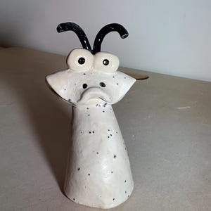 May include: A whimsical ceramic sculpture of a creature with large, round eyes and black antennae. The off-white body is speckled with dark blue spots. The sculpture has a cone-shaped base and a stylized face.