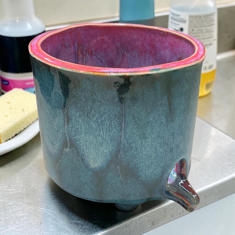 May include: A cylindrical ceramic planter with a glossy finish, featuring a deep teal and dark blue mottled exterior glaze. The interior and the top rim are coated in a vibrant magenta glaze. The base includes a small, protruding drainage spout.