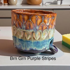 May include: A ceramic bowl with a reactive glaze featuring a gradient of brown, green, and purple colors in a striped pattern. The bowl has a wide, open top and a small, curved spout extending from the lower side. The text Brn Grn Purple Stripes is visible.