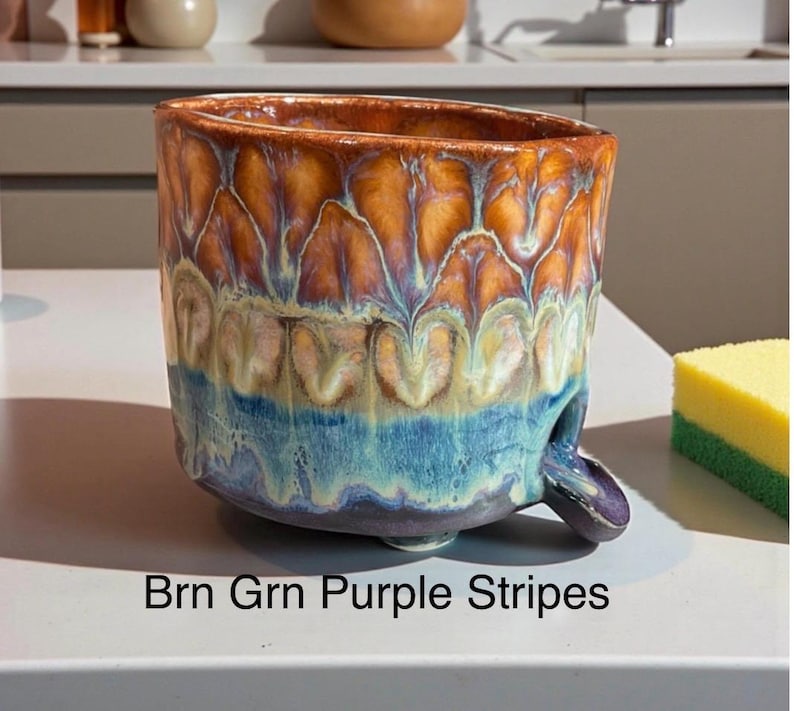 May include: A ceramic bowl with a reactive glaze featuring a gradient of brown, green, and purple colors in a striped pattern. The bowl has a wide, open top and a small, curved spout extending from the lower side. The text Brn Grn Purple Stripes is visible.