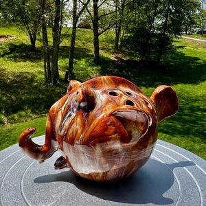 May include: A brown and white ceramic planter shaped like a cartoon pig with a large snout and ears. The planter has a glazed finish and several holes on the top for drainage.