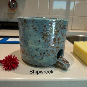 May include: A cylindrical ceramic mug with a light blue glaze and brown speckles throughout. The mug features a small, protruding spout at the base and the word Shipwreck printed in black text below it. It has a textured, hand-formed appearance.