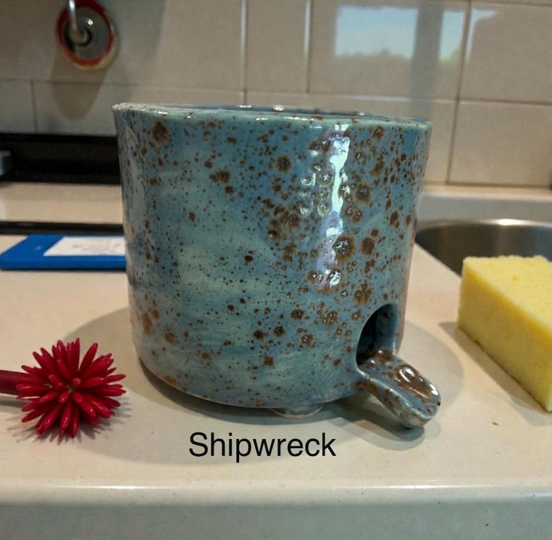 May include: A cylindrical ceramic mug with a light blue glaze and brown speckles throughout. The mug features a small, protruding spout at the base and the word Shipwreck printed in black text below it. It has a textured, hand-formed appearance.