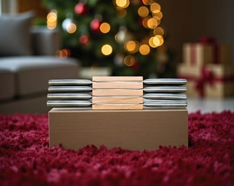 May include: A stack of wooden and metal cheese knives arranged on a brown box, set against a blurred background of a Christmas tree with colorful lights and ornaments. The knives are silver and wood-toned.