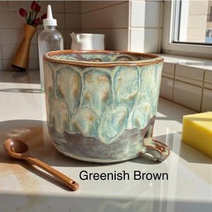 May include: A cylindrical ceramic bowl with a reactive glaze featuring a blend of light green, turquoise, and brown tones. The bowl has a small, protruding spout at the base and a smooth, rounded rim. The text Greenish Brown is visible below.