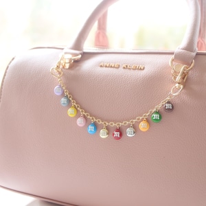 Peut inclure: Un sac à main rose avec une chaîne dorée et des breloques M&M colorées attachées. Le sac à main porte l'inscription "Anne Klein" sur le devant.
