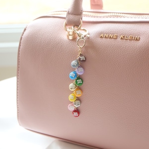 May include: A pink handbag with a gold chain and a charm featuring a rainbow of M&M's. The bag has the text "Anne Klein" on the front.