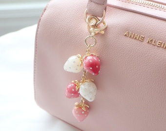 May include: A pink handbag with a gold chain and a charm featuring a cluster of pink and white strawberry-shaped ornaments. The bag has the text "Anne Klein" on the front.