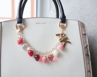May include: A white handbag with a gold chain and strawberry charms. The bag has a black handle and the brand name "Anne Klein" is printed on the front.