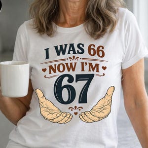 Puede incluir: Camiseta blanca con el texto "I WAS 66 NOW I'M 67" en marr&oacute;n y azul marino. El dise&ntilde;o incluye dos manos y corazones. La persona lleva una taza blanca.