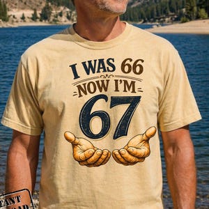 Puede incluir: Camiseta beige de manga corta con el texto "I WAS 66 NOW I'M 67" en marr&oacute;n y azul marino. La camiseta presenta una ilustraci&oacute;n de dos manos abiertas. La camiseta la lleva una persona.