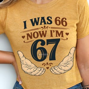 Puede incluir: Camiseta de color amarillo mostaza con el texto "I WAS 66 NOW I'M 67" en azul marino y marr&oacute;n. El n&uacute;mero 67 es grande y azul marino, con dos manos abiertas a cada lado. La camiseta es de estilo crop.