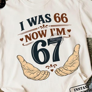 May include: White t-shirt with the text "I WAS 66 NOW I'M 67" in navy blue and brown. The number 67 is in navy blue, with two hands illustrated below. A fun birthday gift for someone turning 67.
