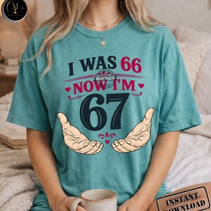 Puede incluir: Camiseta de color turquesa con el texto "I WAS 66 NOW I'M 67" en rojo y negro. El dise&ntilde;o incluye dos manos y el logotipo de Merytouch. Se ve una taza de caf&eacute; en primer plano.