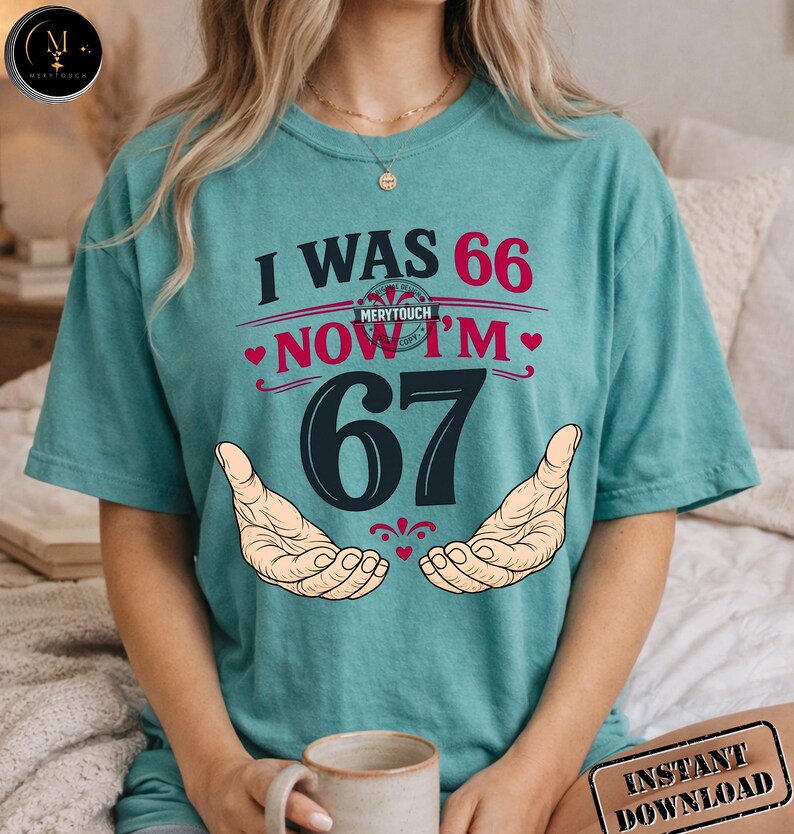 Puede incluir: Camiseta de color turquesa con el texto "I WAS 66 NOW I'M 67" en rojo y negro. El dise&ntilde;o incluye dos manos y el logotipo de Merytouch. Se ve una taza de caf&eacute; en primer plano.