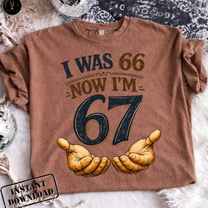 Puede incluir: Una camiseta marr&oacute;n con el texto "I WAS 66 NOW I'M 67" en azul marino y marr&oacute;n. El n&uacute;mero 67 es en azul marino con un contorno marr&oacute;n. Dos manos est&aacute;n ilustradas debajo del n&uacute;mero 67.