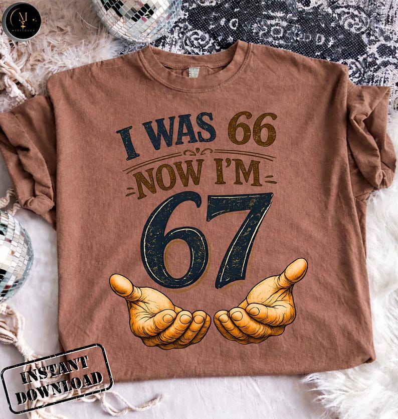 Puede incluir: Una camiseta marr&oacute;n con el texto "I WAS 66 NOW I'M 67" en azul marino y marr&oacute;n. El n&uacute;mero 67 es en azul marino con un contorno marr&oacute;n. Dos manos est&aacute;n ilustradas debajo del n&uacute;mero 67.