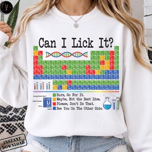 Op de afbeelding: Witte sweatshirt met de tekst "Can I Lick It?" boven een kleurrijk periodiek systeem der elementen. Daaronder staan de zinnen "Sure, Go For It", "Maybe, Not the Best Idea" en "Please, Don't Do That."