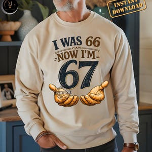 Puede incluir: Sudadera beige con el texto "I WAS 66 NOW I'M 67" en azul marino y marr&oacute;n. El n&uacute;mero 67 es grande y azul oscuro. El dise&ntilde;o incluye ilustraciones de manos. Las palabras "Instant Download" est&aacute;n en la esquina superior derecha.