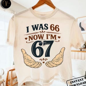 Puede incluir: Camiseta color crema con el texto "I WAS 66 NOW I'M 67" en marr&oacute;n y azul marino. El dise&ntilde;o incluye dos manos abiertas y corazones. La camiseta est&aacute; colgada en una percha de madera.