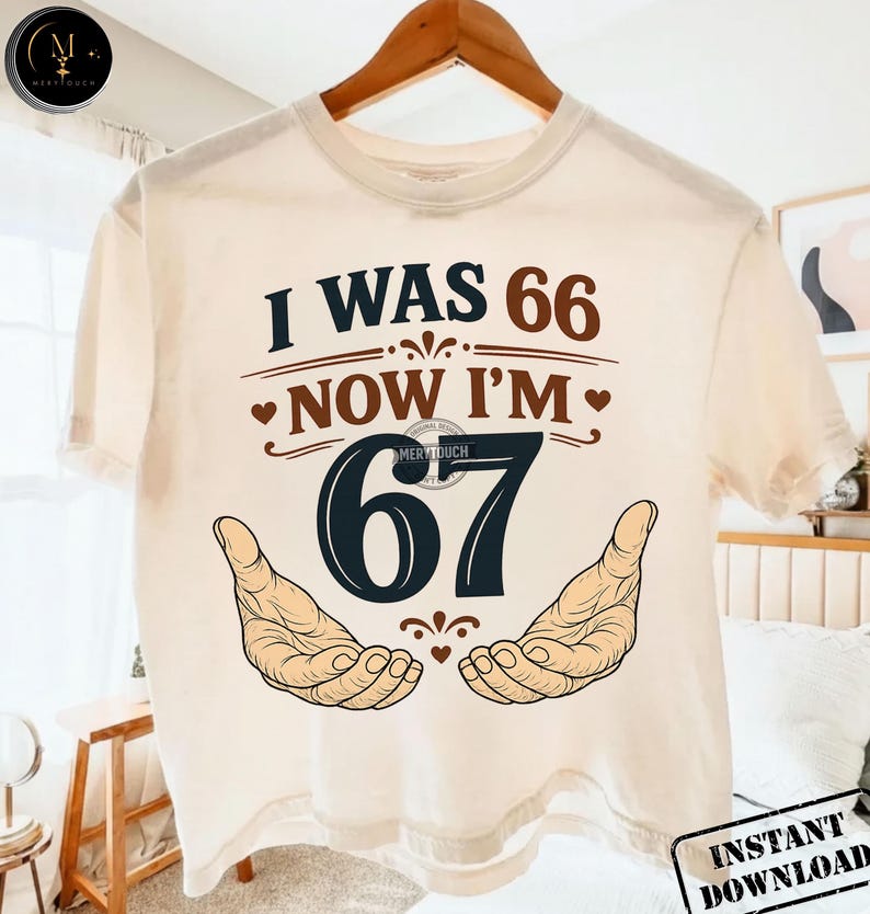 Puede incluir: Camiseta color crema con el texto "I WAS 66 NOW I'M 67" en marr&oacute;n y azul marino. El dise&ntilde;o incluye dos manos abiertas y corazones. La camiseta est&aacute; colgada en una percha de madera.