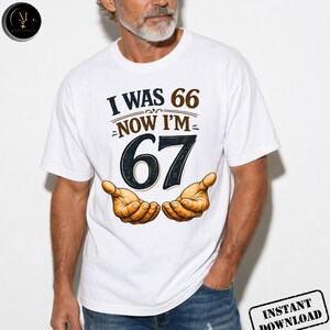 Puede incluir: Camiseta blanca con el texto "I WAS 66 NOW I'M 67" en marr&oacute;n y negro. El n&uacute;mero 67 es grande y est&aacute; centrado. Dos manos abiertas est&aacute;n debajo del texto. La camiseta la lleva una persona.