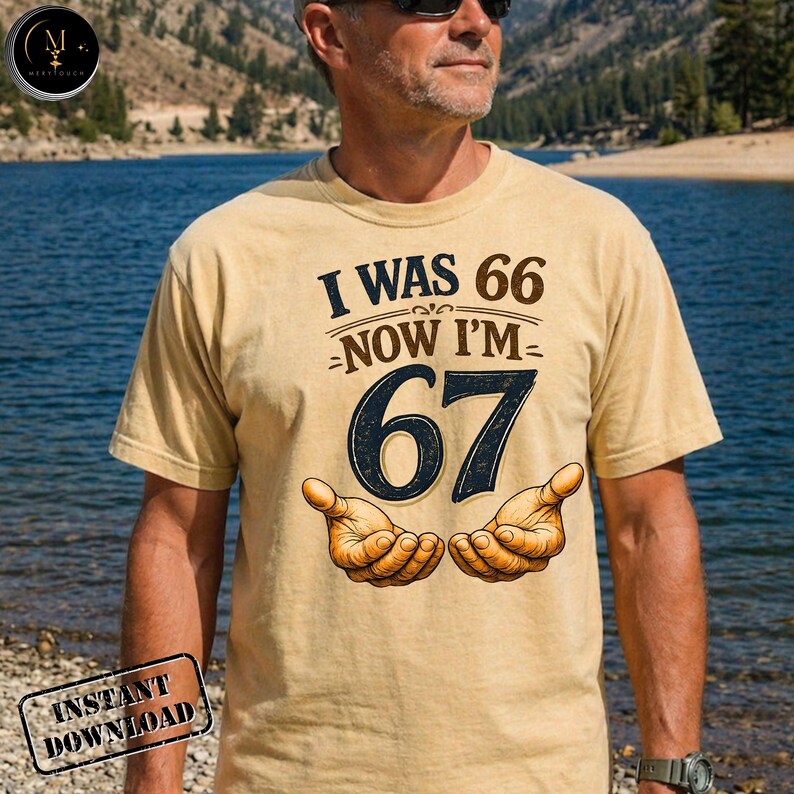 Puede incluir: Camiseta beige de manga corta con el texto "I WAS 66 NOW I'M 67" en marr&oacute;n y azul marino. La camiseta presenta una ilustraci&oacute;n de dos manos abiertas. La camiseta la lleva una persona.