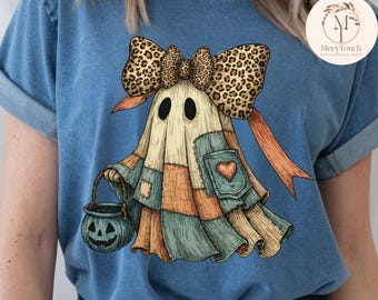 May include: A blue t-shirt featuring a whimsical ghost design. The ghost has a patchwork body, a leopard print bow, and a pumpkin-shaped candy bucket. The design includes a heart-shaped patch and orange ribbons, perfect for Halloween.